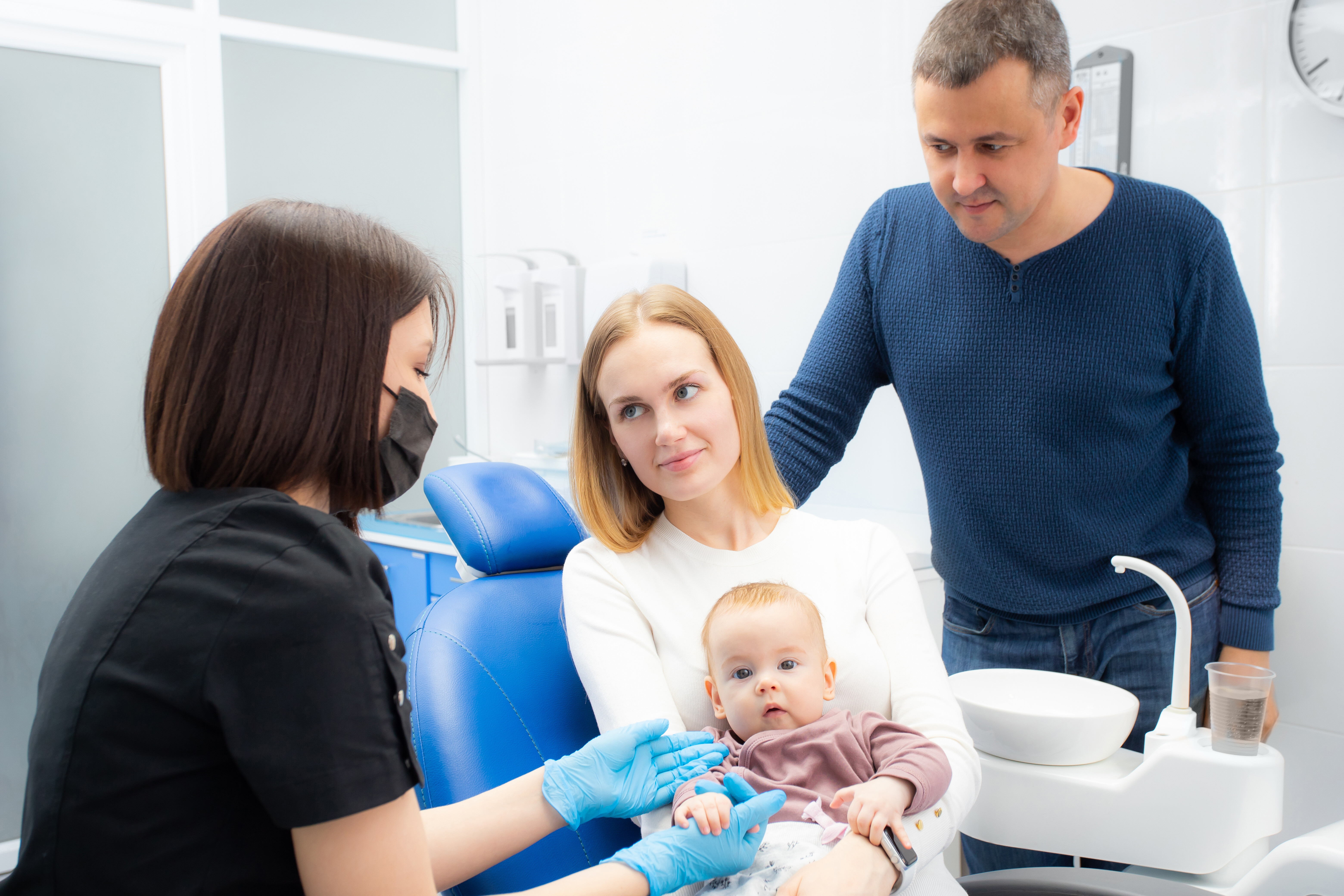 Young parents and their daughter visit a dentist to examine milk teeth. Baby at the first appointment with the dentist. Inspection of the formation of jaws, eruption of milk teeth